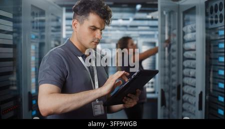 Professional IT Specialist Operates Tablet Computer to Monitor System Status while Female Colleague Inspects Server Rack Hardware. Team Collaboration Ensures Smooth Cloud Infrastructure Performance. Stock Photo