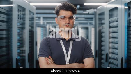 In Modern High Tech Data Center, Male Network Engineer Poses Confidently Between Server Racks. Reliable Hardware Infrastructure and Cybersecurity Systems Protect Enterprise Digital Assets. Portrait. Stock Photo