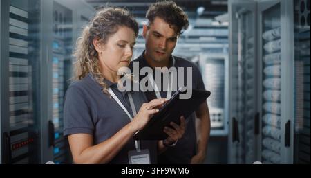 In Modern Data Center, Female Engineer Uses Digital Tablet while Colleague Checks Server Racks. Mobile Cloud Diagnostics and Team Collaboration Ensure High Tech Network Stability. Concept of Teamwork. Stock Photo