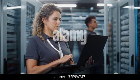 In Modern Data Center Server Room, Female IT Specialist Works on Laptop While Male Colleague Inspects Server Racks. System Administration and Teamwork Ensure Continuous Cloud Uptime. Big Data Concept. Stock Photo