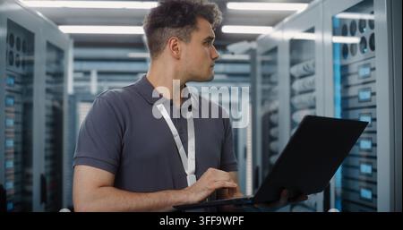 Focused Network Engineer Stands in Server Room, Using Laptop to Configure Enterprise Hardware. Cyber Security and Cloud Storage Management Protect Critical Data Systems. Infrastructure Monitoring. Stock Photo