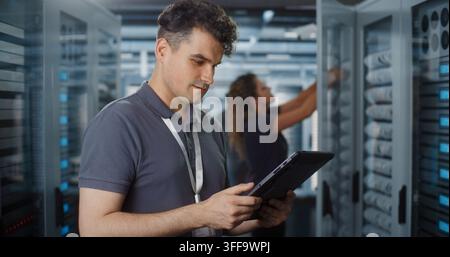 Professional IT Specialist Operates Tablet Computer to Monitor System Status while Female Colleague Inspects Server Rack Hardware. Team Collaboration Ensures Smooth Cloud Infrastructure Performance. Stock Photo