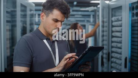 Standing in Server Room, Technician Analyzes Data Metrics on Tablet as Teammate Configures Server Rack Components. Mobile Diagnostic Tools and Physical Maintenance Optimize Cloud Server Efficiency. Stock Photo
