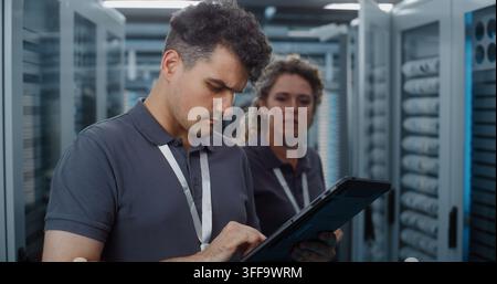 Standing in Server Room, Technician Analyzes Data Metrics on Tablet as Teammate Configures Server Rack Components. Mobile Diagnostic Tools and Physical Maintenance Optimize Cloud Server Efficiency. Stock Photo