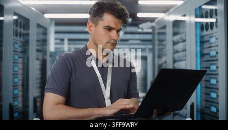 Focused Network Engineer Stands in Server Room, Using Laptop to Configure Enterprise Hardware. Cyber Security and Cloud Storage Management Protect Critical Data Systems. Infrastructure Monitoring. Stock Photo