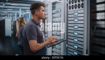Focused Network Technician Holds Tablet Device while Verifying Server Rack Labels and Hardware Setup. Mobile Diagnostics and Structured Cabling Management Maintain Enterprise Cloud Performance. Stock Photo
