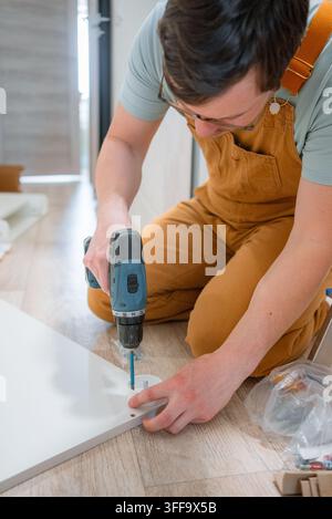 Man wearing overalls and glasses assembling new furniture, using an electric power drill to screw components for a chest of drawers onto a light wood floor during a home renovation project Stock Photo