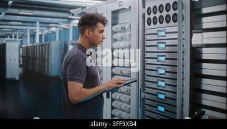 IT Engineer Holds Digital Tablet while Touching and Inspecting Server Rack Modules in Modern Data Center. Routine Hardware Maintenance, Physical Checks Ensure High Availability Server Infrastructure. Stock Photo