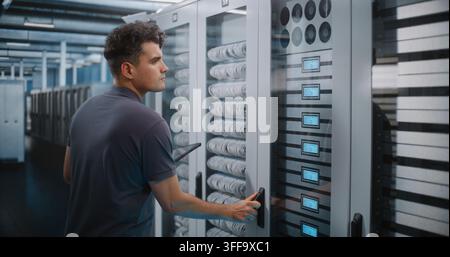 Focused Network Technician Holds Tablet Device while Verifying Server Rack Labels and Hardware Setup. Mobile Diagnostics and Structured Cabling Management Maintain Enterprise Cloud Performance. Stock Photo