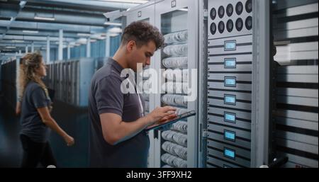 IT Engineer Holds Digital Tablet while Touching and Inspecting Server Rack Modules in Modern Data Center. Routine Hardware Maintenance, Physical Checks Ensure High Availability Server Infrastructure. Stock Photo