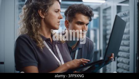 Focused Female System Administrator Types on Laptop While Colleague Conducts Server Cabinet Check in Data Center Corridor. Women in STEM Drive Modern Enterprise IT Operations and Cloud Security. Stock Photo
