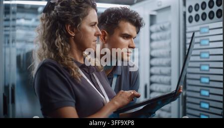 Focused Female System Administrator Types on Laptop While Colleague Conducts Server Cabinet Check in Data Center Corridor. Women in STEM Drive Modern Enterprise IT Operations and Cloud Security. Stock Photo