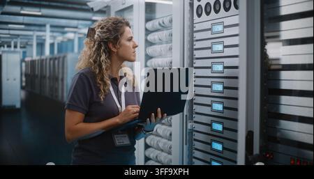 IT Specialist Holds Laptop Computer to Inspect Server Racks and Configure Cloud Infrastructure in Data Center Server Room. System Diagnostics and Hardware Maintenance Ensure Seamless Network Uptime. Stock Photo