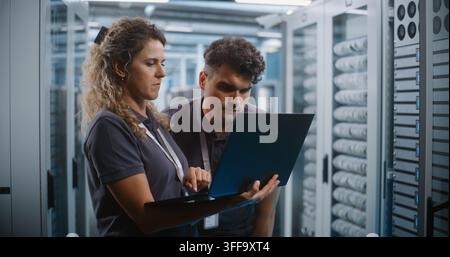 In Server Room Corridor, Two Network Engineers Inspect Cybersecurity Metrics and System Logs on Laptop Screen. Joint Diagnostics and Enterprise Cloud Monitoring Protect Critical Server Networks. Stock Photo