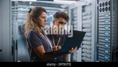 In Server Room Corridor, Two Network Engineers Inspect Cybersecurity Metrics and System Logs on Laptop Screen. Joint Diagnostics and Enterprise Cloud Monitoring Protect Critical Server Networks. Stock Photo