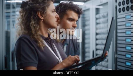 Focused Female System Administrator Types on Laptop While Colleague Conducts Server Cabinet Check in Data Center Corridor. Women in STEM Drive Modern Enterprise IT Operations and Cloud Security. Stock Photo