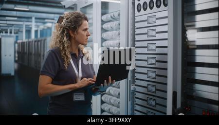 IT Specialist Holds Laptop Computer to Inspect Server Racks and Configure Cloud Infrastructure in Data Center Server Room. System Diagnostics and Hardware Maintenance Ensure Seamless Network Uptime. Stock Photo