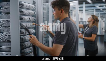 In Data Center, Male Technician Vacuums Dust From Server Cables With Brush While Female Colleague Cleans Cabinet Glass. Professional IT Facility Maintenance Guarantees Enterprise Server Reliability. Stock Photo