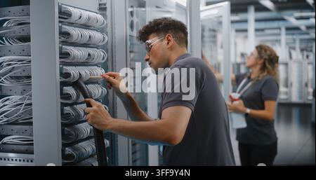 In Data Center, Male Technician Vacuums Dust From Server Cables With Brush While Female Colleague Cleans Cabinet Glass. Professional IT Facility Maintenance Guarantees Enterprise Server Reliability. Stock Photo