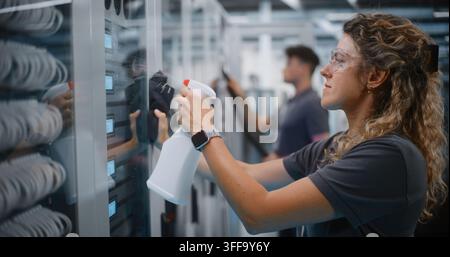IT Maintenance Team Works in High Tech Server Corridor. Female Technician Sanitizes Glass Rack Doors With Spray Cleaner While Male Technician Operates Vacuum. Comprehensive Facility Upkeep Concept. Stock Photo