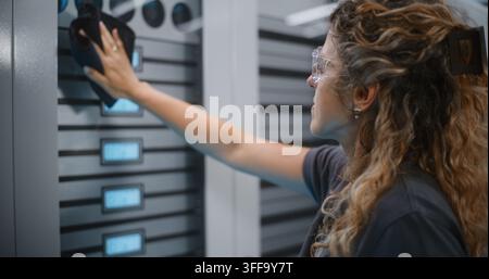 Female System Specialist in Safety Goggles Carefully Cleans Glass Server Rack Door With Cloth and Cleaning Spray. Women in Technology Maintain Enterprise Server Hygiene and Cloud Network Performance. Stock Photo