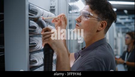 Focused Network Administrator Wearing Safety Glasses Carefully Removes Dust From Server Rack Cables. Preventive Dust Control Safeguards Enterprise Cloud Storage Infrastructure Against Overheating. Stock Photo