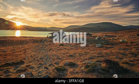 Enontekio, Finland - 28.08.2026: Isolated timber hut and camping tent on rocky tundra terrain with a glowing sun reflecting on a calm lake Stock Photo