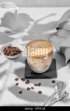 Dalgona coffee. Whipped instant coffee in a glass with ice on a slate board on a gray background with coffee beans  and shadow. The concept of a trend Stock Photo