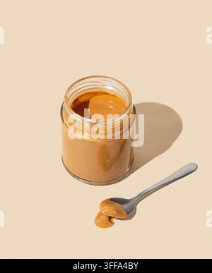 Peanut butter or caramel sauce in glass jar on isolated beige background with spoon and shadow. Concept of tasty and healthy snack for breakfast. Top Stock Photo