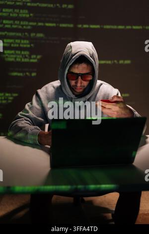 Vertical portrait of dangerous hooded hacker male in sunglasses using smartphone and holding red credit card, surrounded by green code visuals suggest Stock Photo