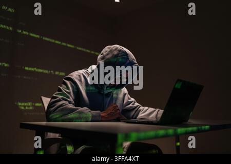 Hooded hacker male in red sunglasses typing on laptop keyboard amid glowing green code on dark walls and table, evoking cybercrime, data theft and int Stock Photo