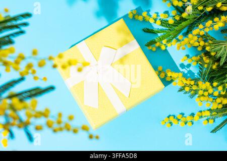 Yellow gift box on blue background with mimosa flowers and sunlight. Concept of advertising banner or greeting card for Mother's Day or spring holiday Stock Photo
