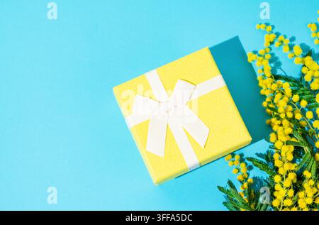 Yellow gift box on blue background with mimosa flowers and sunlight. Concept of advertising banner or greeting card for Mother's Day or spring holiday Stock Photo