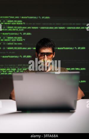 Portrait of skilled young man wearing distinctive sunglasses and developing secret software using laptop, green digital code reflecting on face and t- Stock Photo
