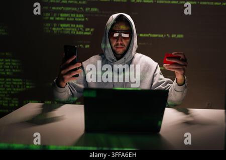 Front view of hooded fraudster in glasses sitting at desk staring at smartphone while holding credit card, laptop open and green code reflecting on wa Stock Photo