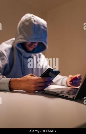 Vertical shot of hooded male engaging in cybercrime, performing online fraud and data theft using mobile phone, laptop and credit card in dark environ Stock Photo