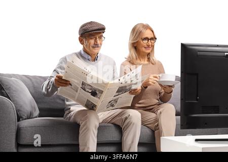 Elderly couple sitting on a sofa with a newspaper and coffee in front of tv isolated on white background Stock Photo