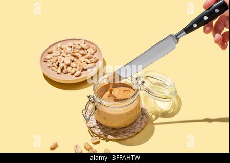 Peanut butter in glass jar on yellow background with knife in hand and salted nuts. Concept of tasty and healthy snack for sweet sandwiches for breakf Stock Photo