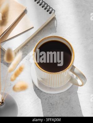 Freshly brewed espresso coffee in a mug on a light table illuminated by the sun and shadow, with notebooks and dried flowers. The concept of a calm mo Stock Photo