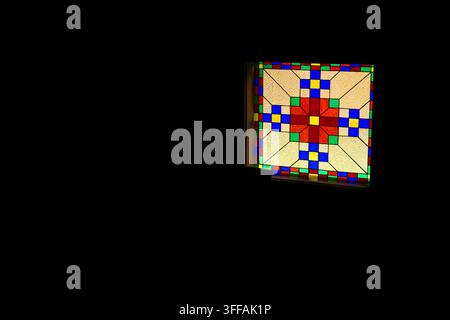 A colorful stained glass window glows against deep darkness showcasing a geometric star motif and vivid tiles that create a focused beacon of light wi Stock Photo