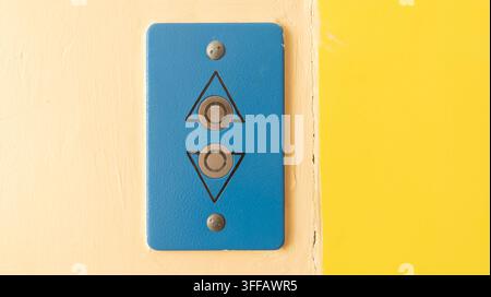 Vintage blue metal elevator call panel. Surface-mounted on a peach and yellow wall, the rectangular faceplate features two silver buttons with engrave Stock Photo