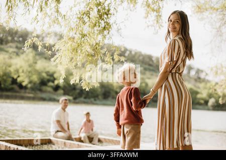 A woman and a child share a warm smile by the riverbank, as two men enjoy a peaceful time in a boat. Sunlight filters through the trees. Stock Photo
