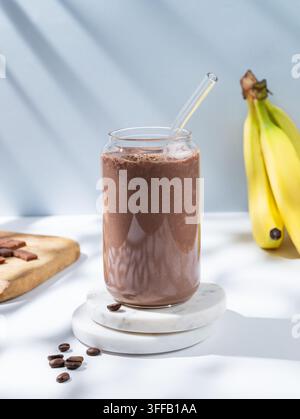 Chocolate coffee smoothie with banana in a glass on a white blue background with morning shadow. Healthy eating and detox diet concept for advertising Stock Photo