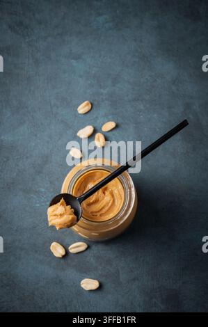 Creamy peanut butter in a jar on a dark background with roasted nuts and spoon close up. Concept for a natural, nutritious vegetarian product. Top vie Stock Photo