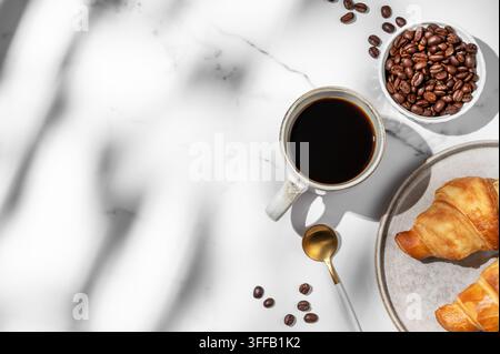 Freshly brewed black coffee in a cup on a white marble background with croissants, coffee beans, spoon and natural morning sunlight. Concept of tasty Stock Photo