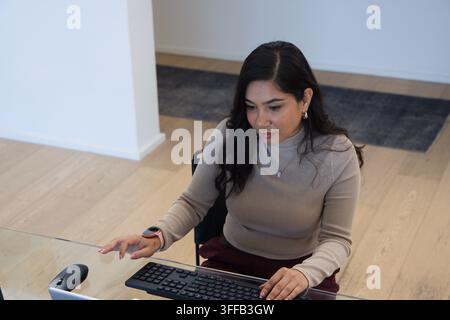 Adult woman typing on keyboard at glass desk in modern workspace, wearing beige ribbed top Stock Photo