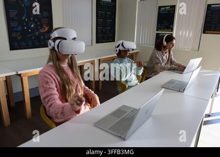Three teenage students wearing white VR headsets while using silver laptops at lab table Stock Photo