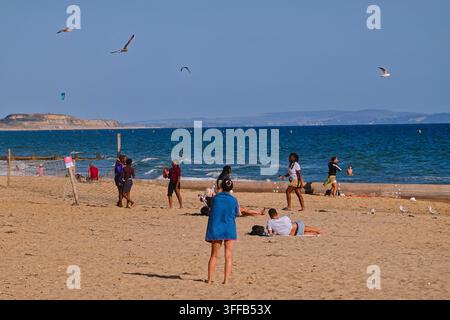 People enjoying a sunny day on the sandy beach at Boscombe, Bournemouth, UK, with seagulls flying over the blue sea. Stock Photo