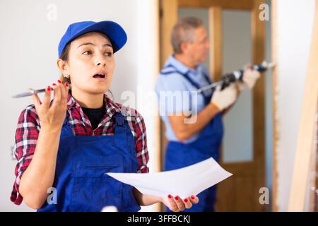 Worried Latin female repairer in a blue overalls calling on a mobile phone while holding a work plan while renovating a house Stock Photo