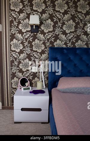 Bedroom bedside scene with patterned beige floral wallpaper, square wall lamp with white shade, deep blue tufted headboard Stock Photo